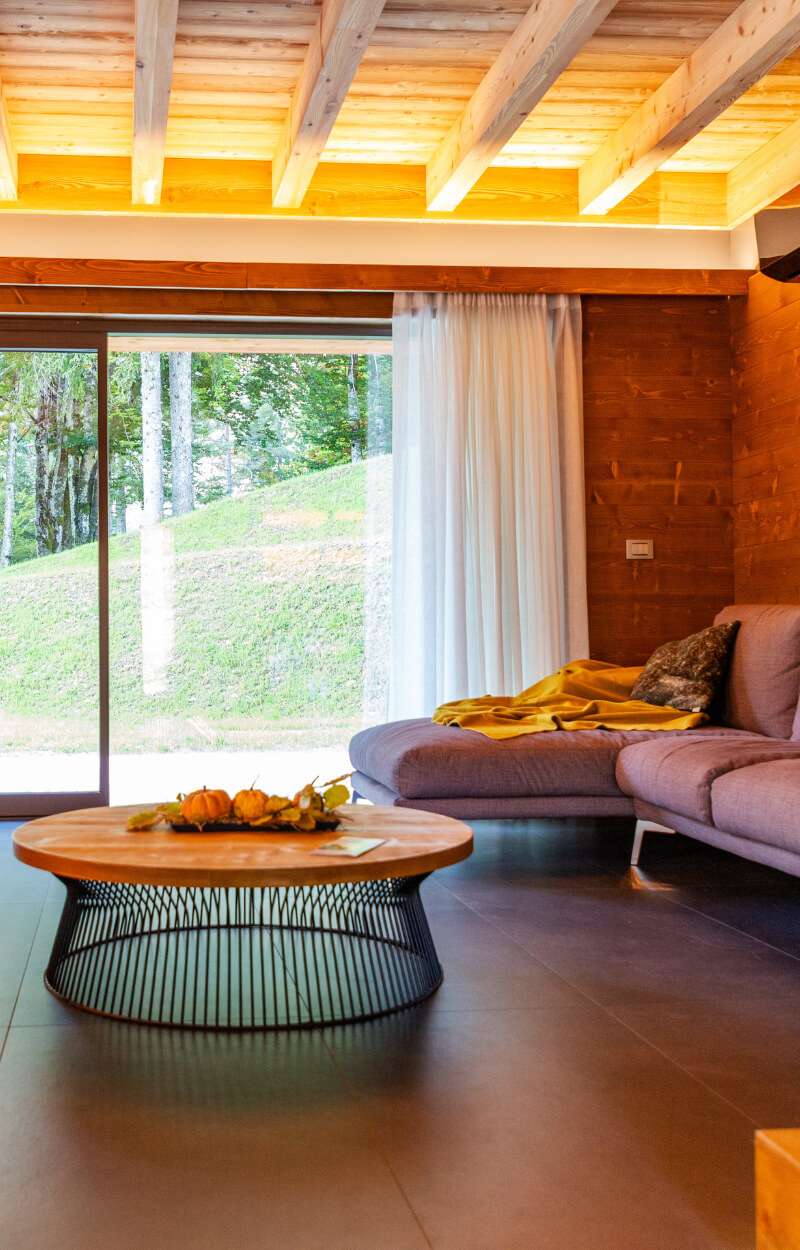 Dnevna soba s pogledom na vrt Chalet Nabois alpskega letovišča Kile v Valbruni Soggiorno con divano e tavolino e vista giardino dello Chalet Nabois del Kile Alpine Resort a Valbruna nella Val Saisera in provincia di Udine